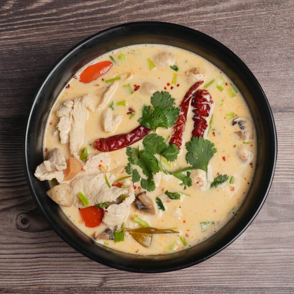 Creamy Tom Kha Gai soup with chicken, mushrooms, red chilies, and cilantro in a black bowl.