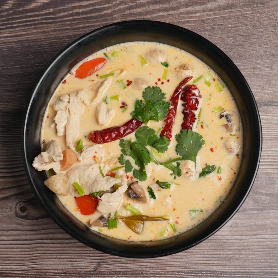 Creamy Tom Kha Gai soup with chicken, mushrooms, red chilies, and cilantro in a black bowl.