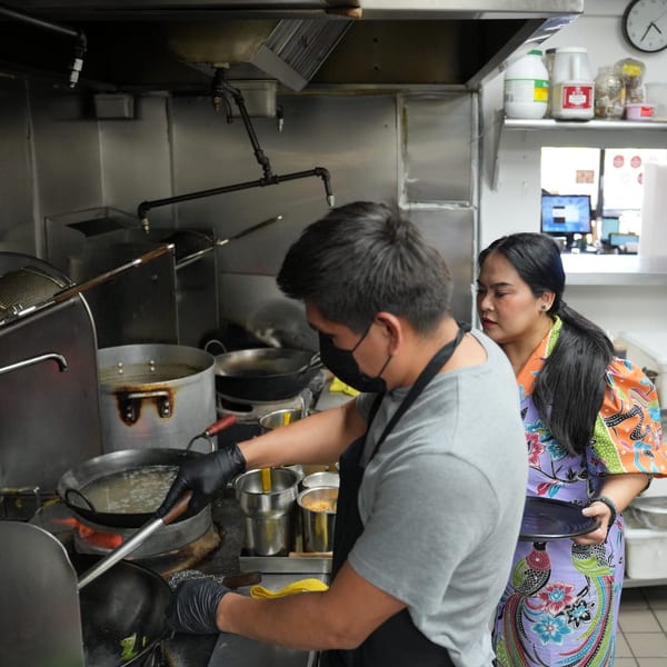 Two Luv2eat Thai Bistro chefs cooking in a busy kitchen, one stirring a wok.