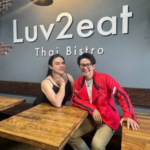 Two smiling people, one in a red jacket, sitting at a wooden table under the Luv2eat Thai Bistro sign.