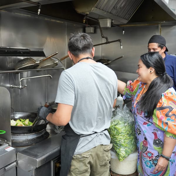 A chef stir-fries food in a wok while a woman in a colorful dress smiles beside him.