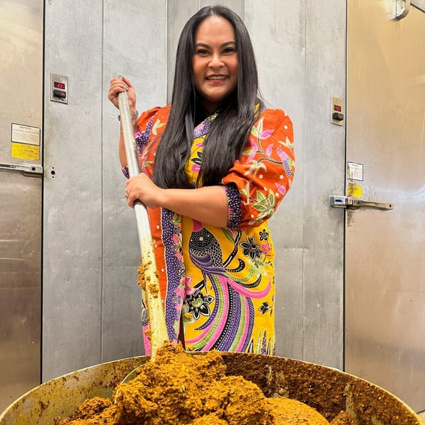 A smiling woman in a colorful dress stirs a large vat of Thai curry paste.