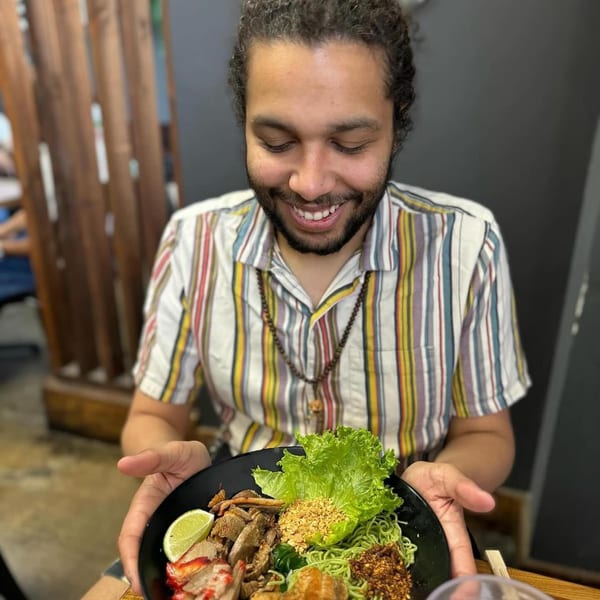 A smiling man in a striped shirt holds a bowl of Luv2eat Thai Bistro's green noodles with meat and lime.
